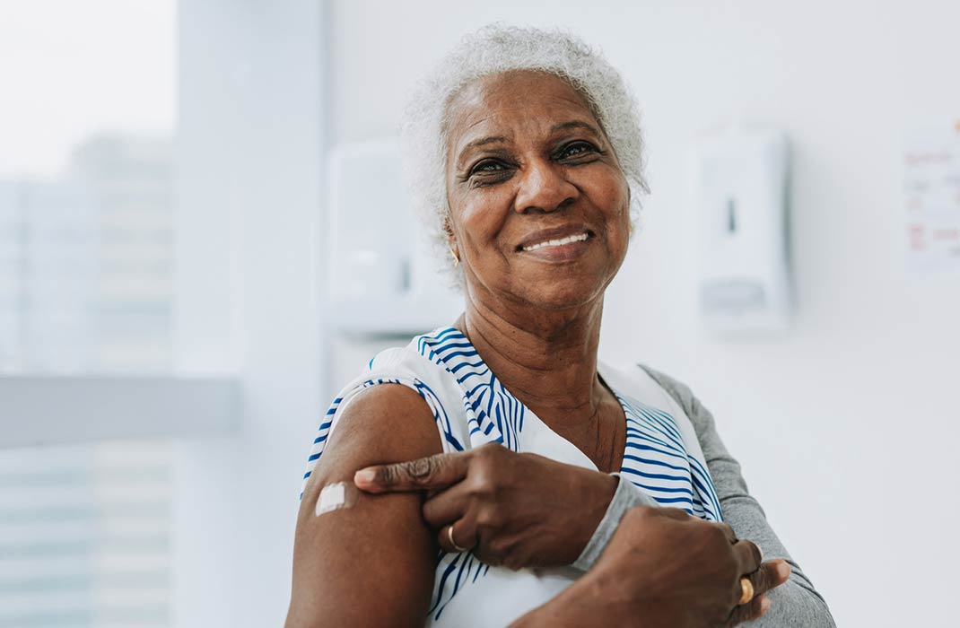 An older adult smiles and points to a bandaid on their right arm after receiving a flu vaccine