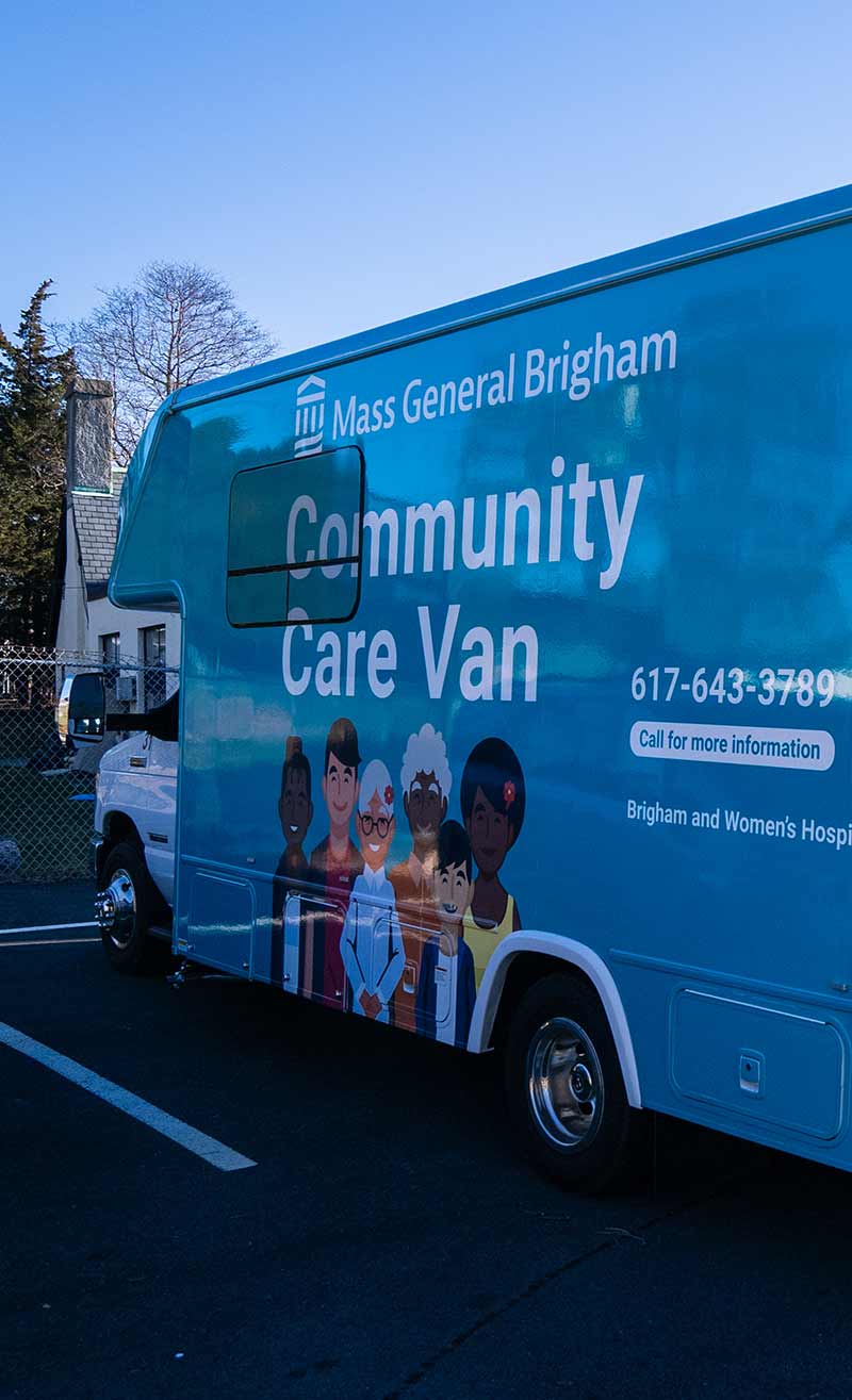 A side view of the Mass General Brigham Community Care Van parked at a location.The van is wrapped in teal on the side and blue on the back consistent with the Mass General Brigham brand colors. The side panel has the Mass General Brigham logo on top with the words "Community Care Van" in large font. Part of the side wrap design includes 5 illustrated individuals of varying genders, ethnicities, and ages.