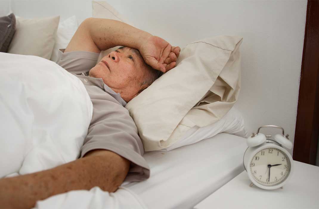A distressed older man lying awake in bed with a clock beside him reading 2:30 am.