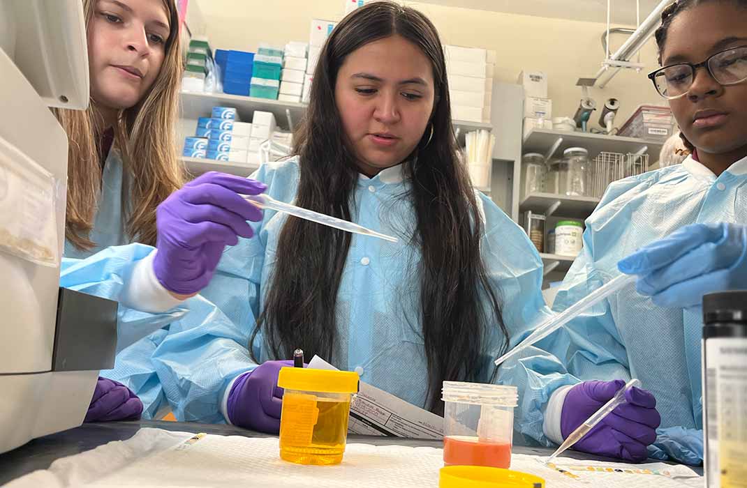 Three individuals wearing protective lab coats and gloves are working in a laboratory setting. They are handling pipettes and sample containers, including a yellow and orange liquid. The background features shelves stocked with laboratory supplies and equipment. 