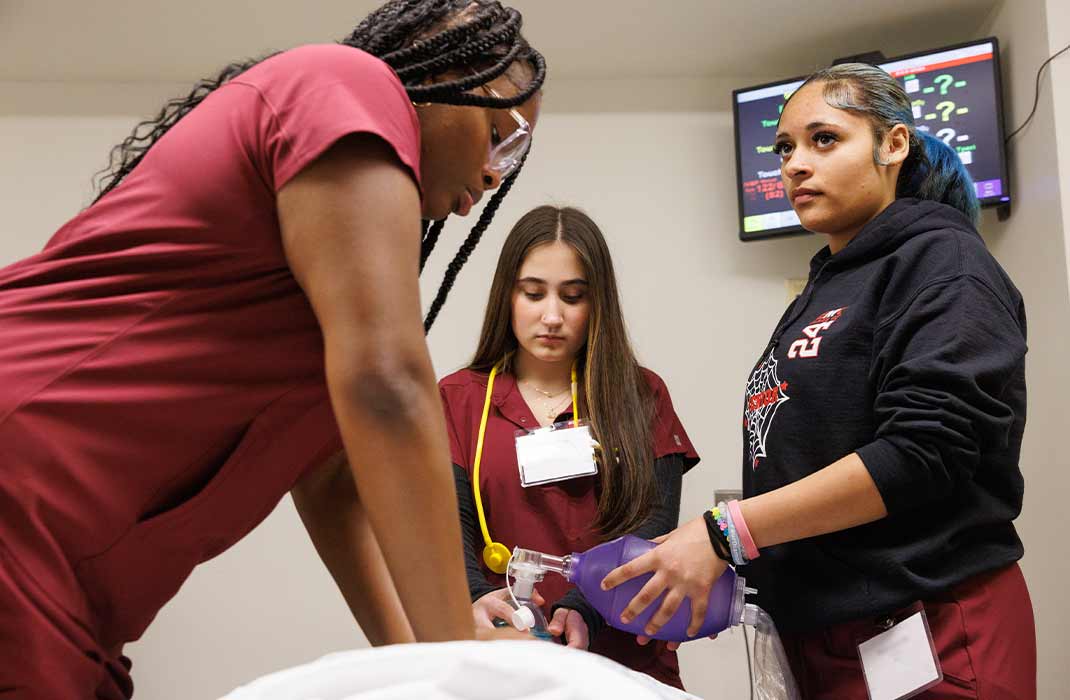 Three students in maroon scrubs practicing in a health care training simulation
