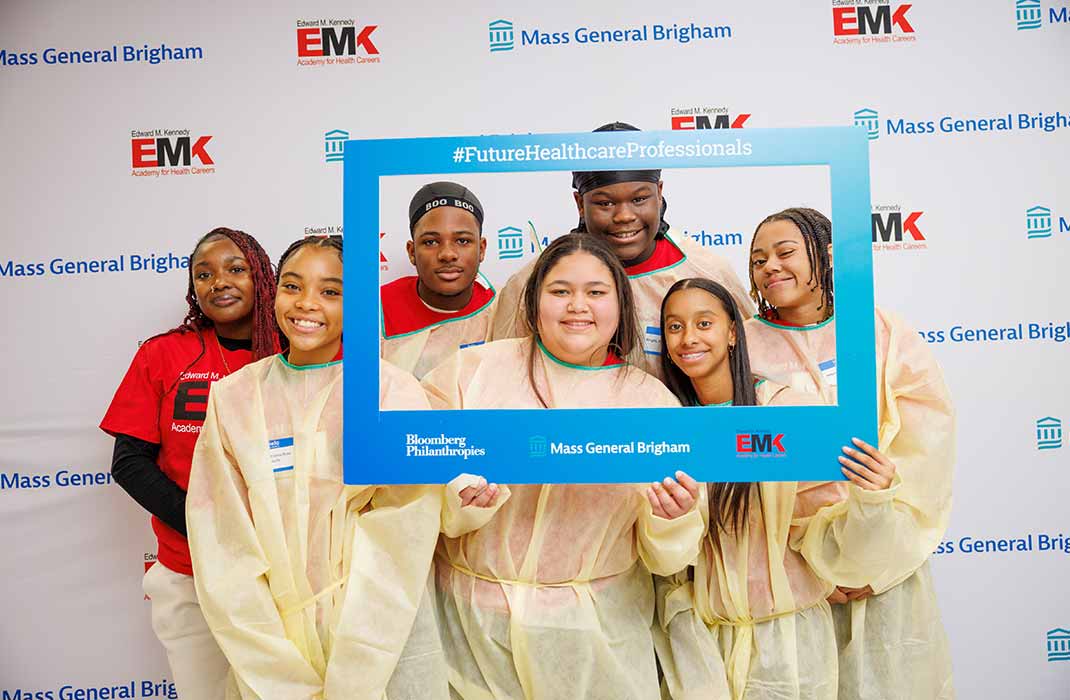 Seven high school students smile and pose with a branded photo frame featuring the logos of Bloomberg Philanthropies, Mass General Brigham, and the Edward M.Kennedy Academy For Health Careers