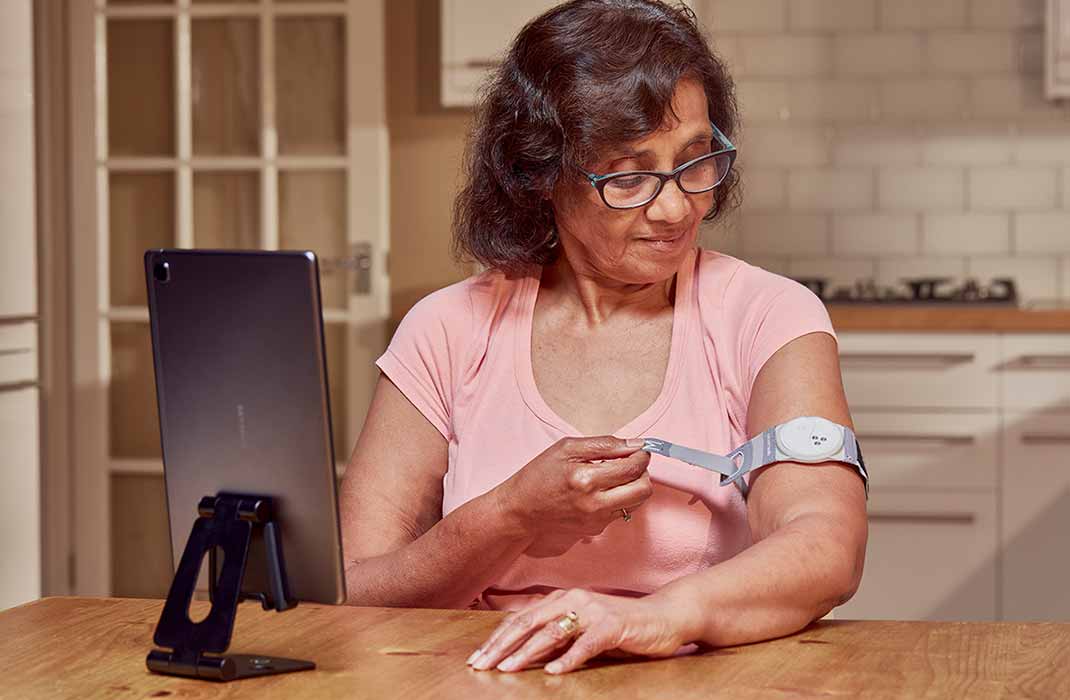 An older woman patient with brown hair, glasses, and a light pink top adjusts her health armband with a tablet in front of her