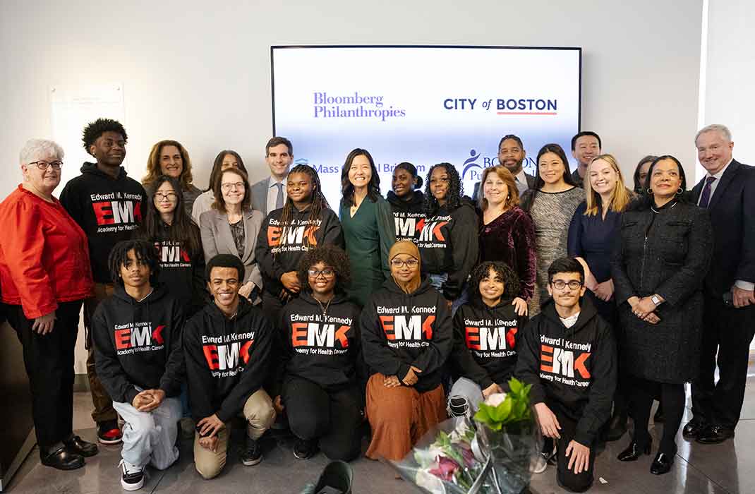 A group of Edward M. Kennedy Academy students in black t-shirts, with Mayor Michelle Wu, Dr. Anne Klibanski, and other leaders and representatives. There is a monitor behind the group with a slide featuring the logos of Bloomberg Philanthropies and the City of Boston.