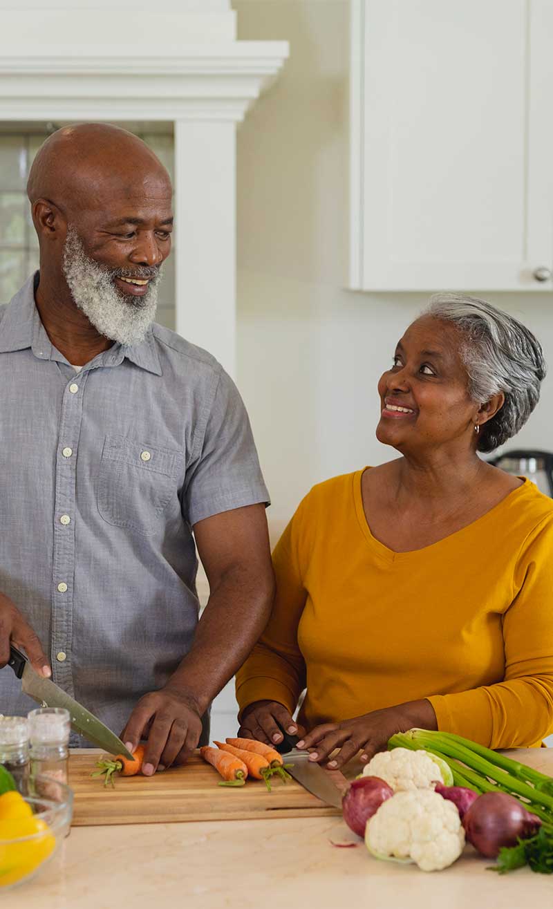 An older couple smile at each other as they chop fresh vegetables for a meal.