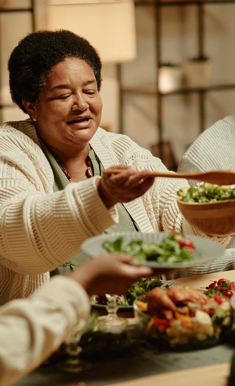 Portrait of smiling African-American grandmother serving food while celebrating Thanksgiving with big happy family at dinner table.