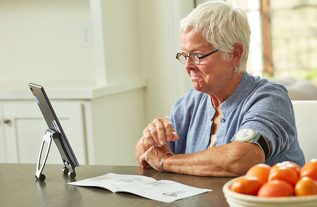 Older woman sits at table, looking at her tablet and wearing a monitor on her arm