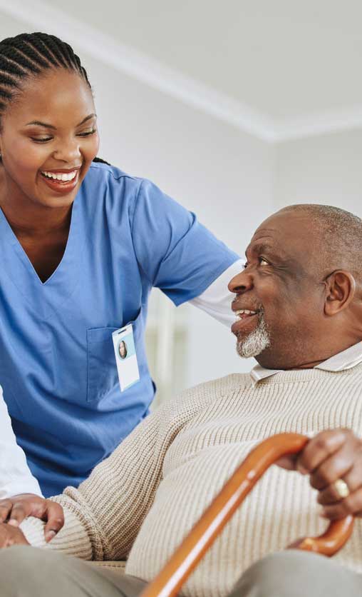 A Black healthcare worker wearing a white long sleeve top under blue scrubs and cornrow braids smiles and checks in on a senior Black man in a beige sweater who is smiling and sitting on a couch while holding a brown cane with his left hand.