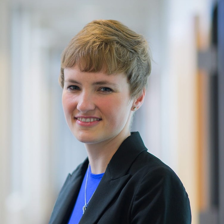 A professionally dressed woman stands in a bright indoor corridor, looking at the camera with a smile. She wears a dark blazer over a blue top, complemented by a simple necklace.