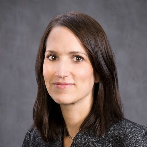 A professional headshot of a woman with shoulder-length dark hair against a neutral gray studio background. She wears a dark textured blazer and a necklace.