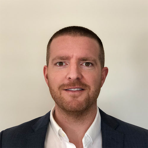 A professional headshot of a man with short hair wearing a white shirt and blazer