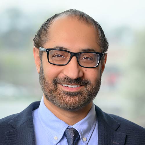 A business professional is shown in a formal portrait wearing a dark suit jacket, light blue dress shirt, a patterned tie, and glasses.