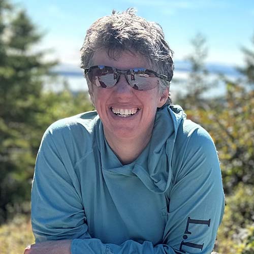 A person wearing a light blue hoodie is seated outdoors, surrounded by greenery and trees.