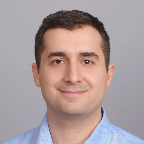 Studio-style portrait headshot of a man with short dark hair wearing a light blue collared shirt against a plain gray background.