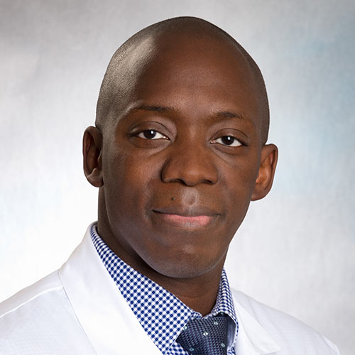 A portrait of an adult male wearing a white lab coat and a blue checkered shirt with a tie.