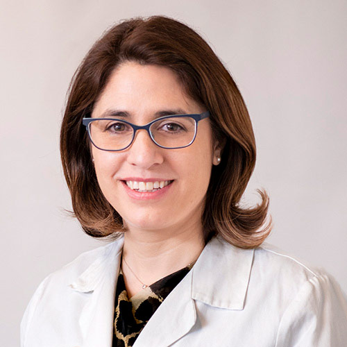 Portrait of a smiling woman wearing glasses and a patterned top under a white medical coat.