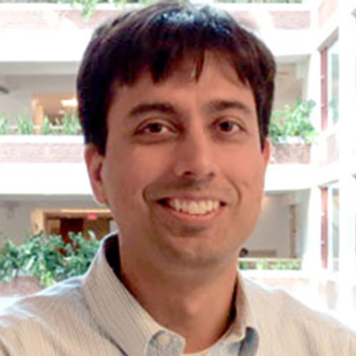 Portrait of a smiling adult man in a light-colored collared shirt. He is standing in an indoor atrium with multiple levels, railings, and green plants visible in the background.