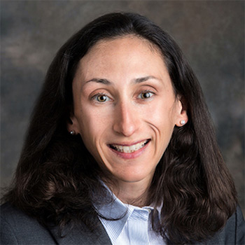 A woman in formal business attire, wearing a gray blazer and light-colored collared shirt, is smiling in her headshot.