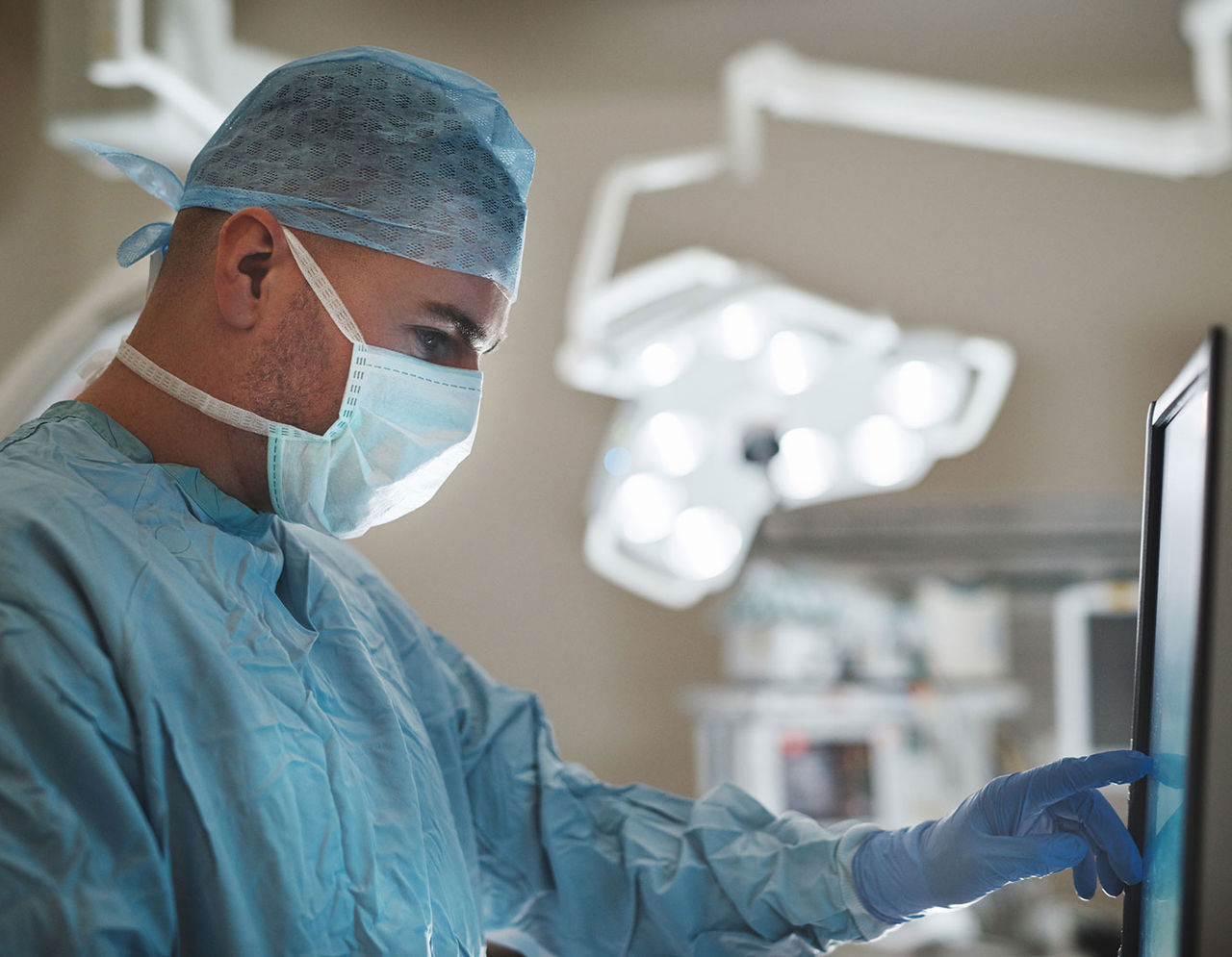 A masked and gowned surgeon uses a touchscreen in the operating room.
