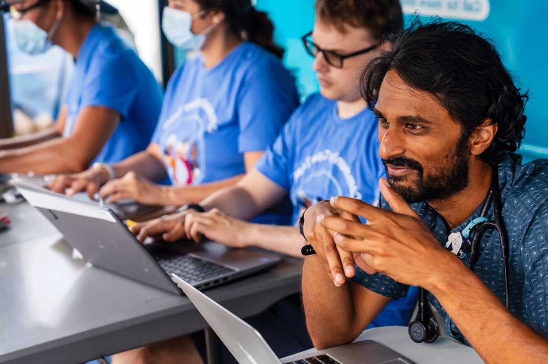 Health care providers in blue Community Care Van T-shirts wait for patients at a van stop.