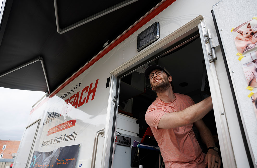 A smiling young man leans out the door of a large clinic van.