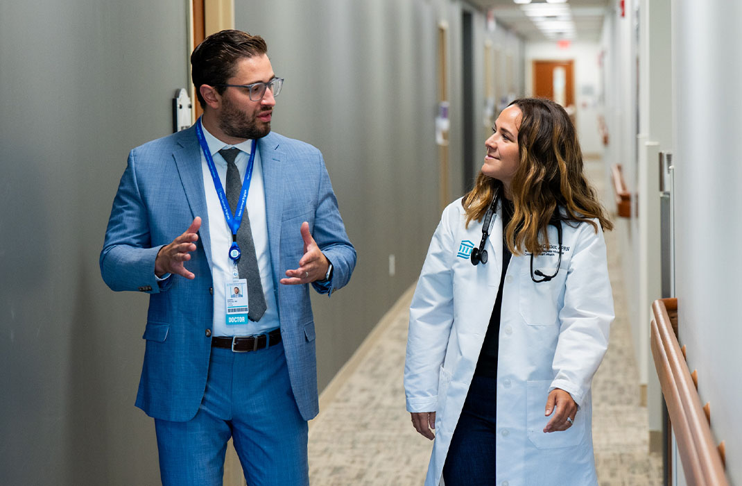 Two clincians are walking and conversing in a hospital corridor. 