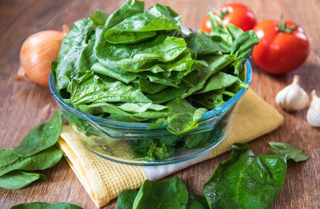 A glass bowl of spinach on a towel that is set on a wooden surface and is surrounded by tomatoes, garlic, and a vidalia onion