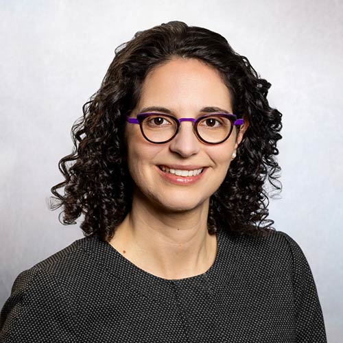 A person with dark, curly hair wearing a textured, dark-colored top with a subtle pattern and eyeglasses is photographed against a neutral, light gray studio background.