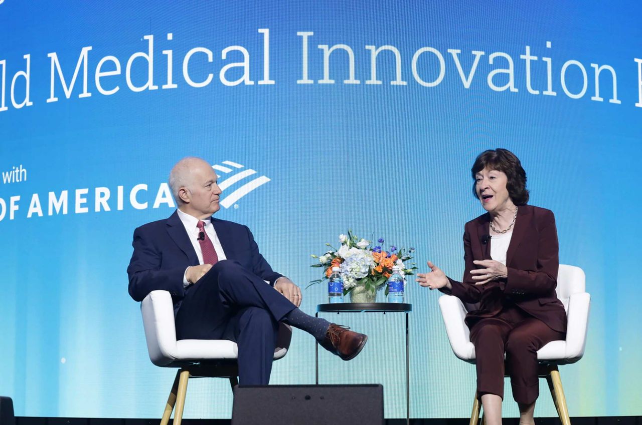 Two individuals seated on stage engage in a discussion during a medical innovation event. The backdrop displays 'World Medical Innovation Forum' and 'Bank of America' text. A floral arrangement is placed on a small table between them.