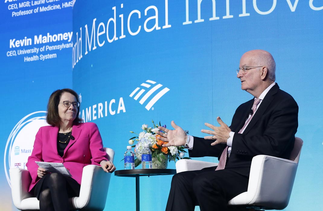 Two individuals are seated on a stage discussing medical innovation. The setting includes a blue backdrop with visible text such as 'World Medical Innovation'. A small table with water bottles and a floral arrangement is placed between them.