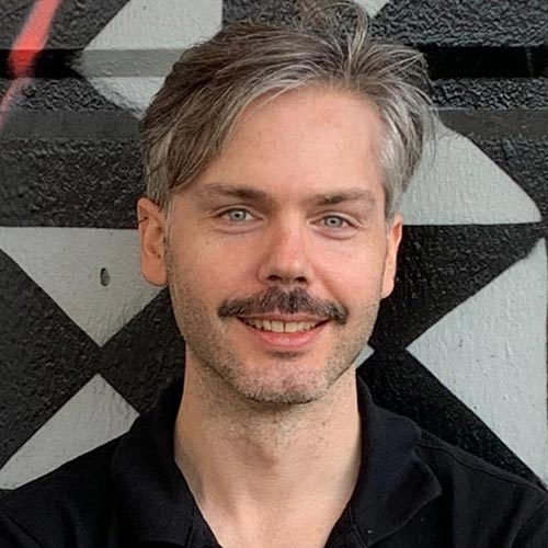 A person wearing a black shirt is seated against a wall with a bold geometric black-and-white pattern.