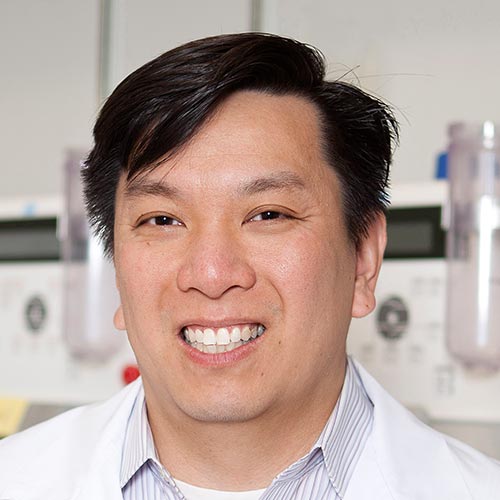 A person wearing a white lab coat is in a laboratory environment. The background features scientific equipment, including monitors and containers.