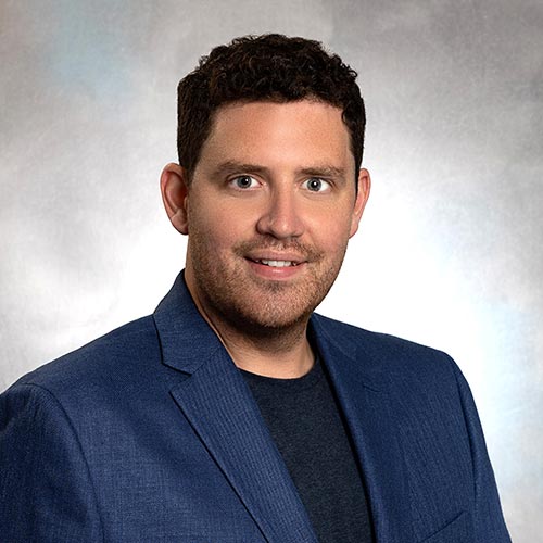 A professional portrait of a man wearing a blue blazer and a dark shirt. The background is a neutral gradient.
