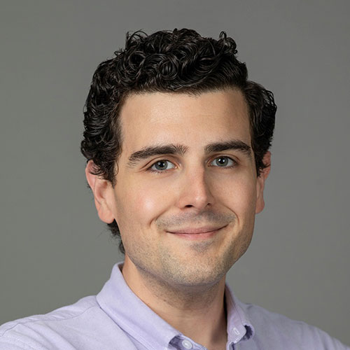 A person with curly dark hair wearing a light purple formal shirt is shown against a neutral gray background.