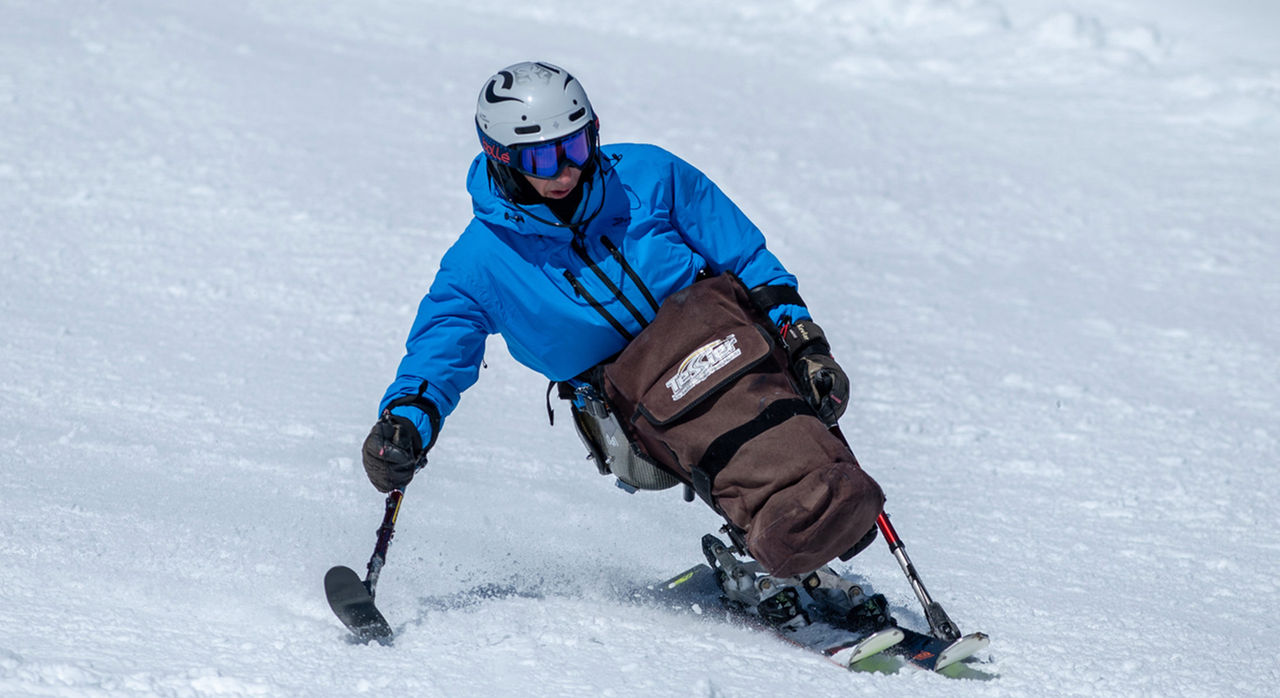 An adaptive skier wearing a blue jacket and helmet navigates a snowy slope using specialized skiing equipment. The skier is focused and in motion.