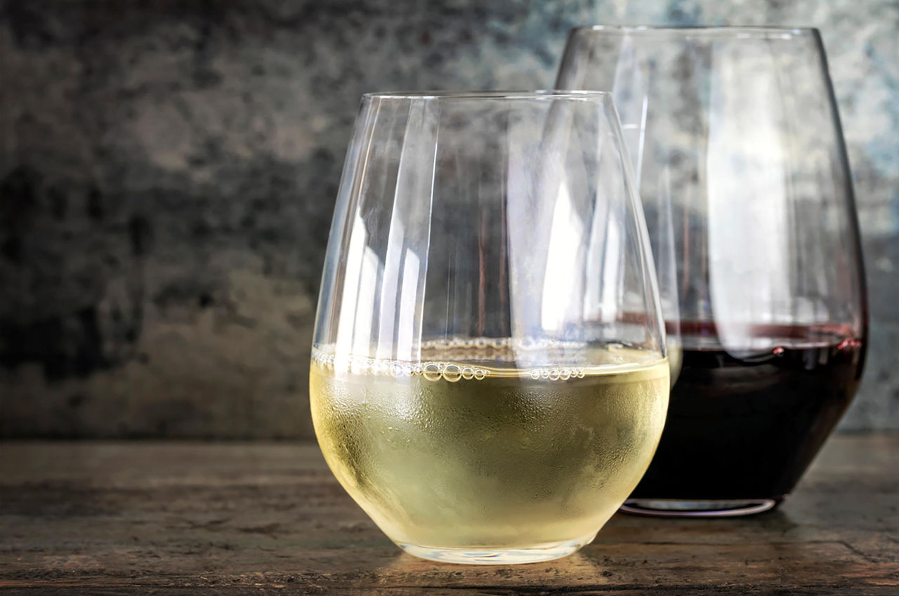 A close-up view of two stemless wine glasses, one filled with white wine and the other with red wine. The glasses are placed on a rustic wooden surface with a textured, neutral background.