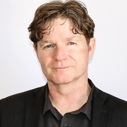 A man is pictured wearing a black blazer over a dark shirt, standing in front of a plain white backdrop.