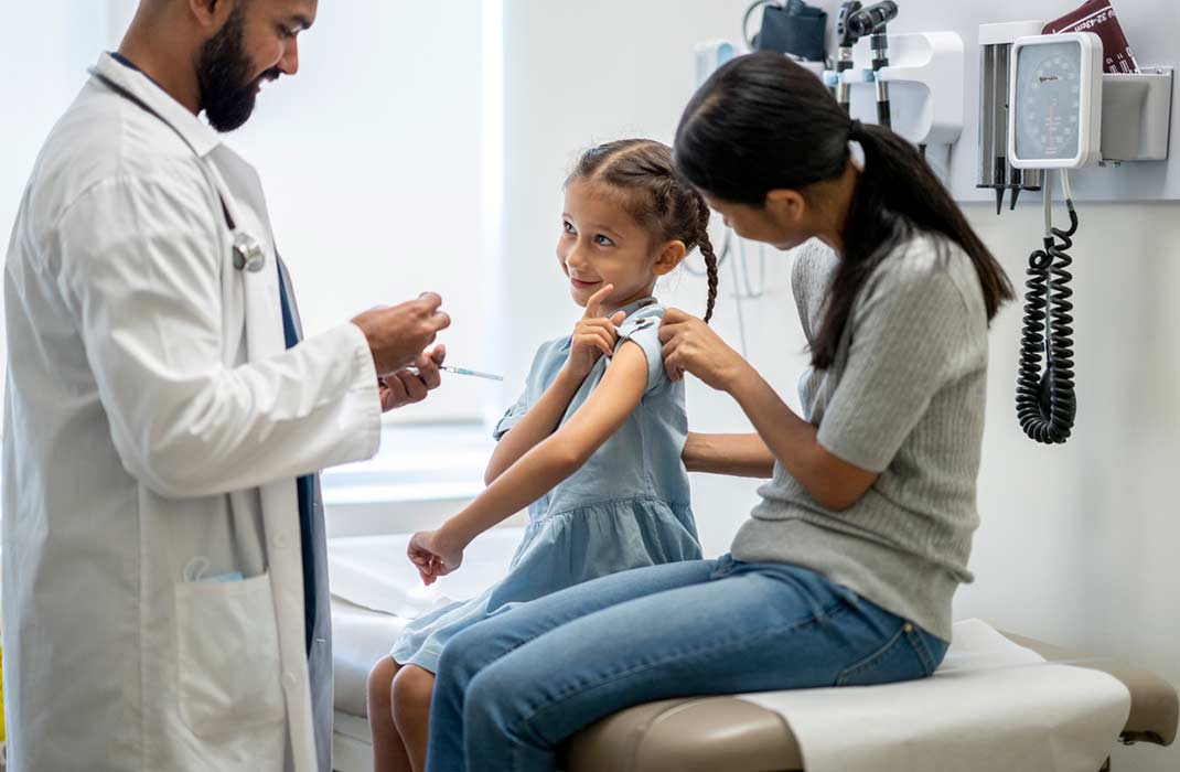 Child receives a flu vaccine from the doctor while their parent sits next to them on an examination table in a doctor's office