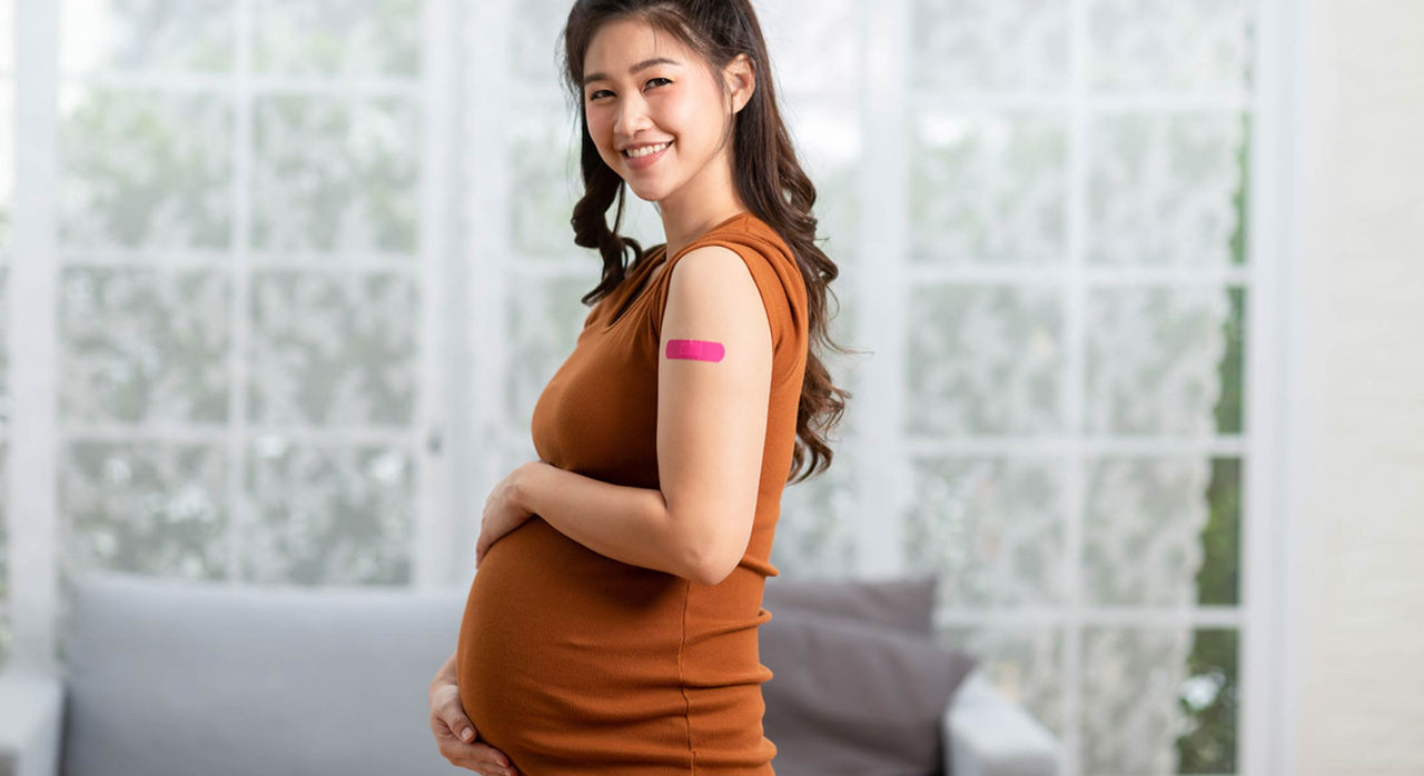 A pregnant patient smiles and holds her baby bump while showing a pink bandaid on her left arm after receiving a flu vaccine