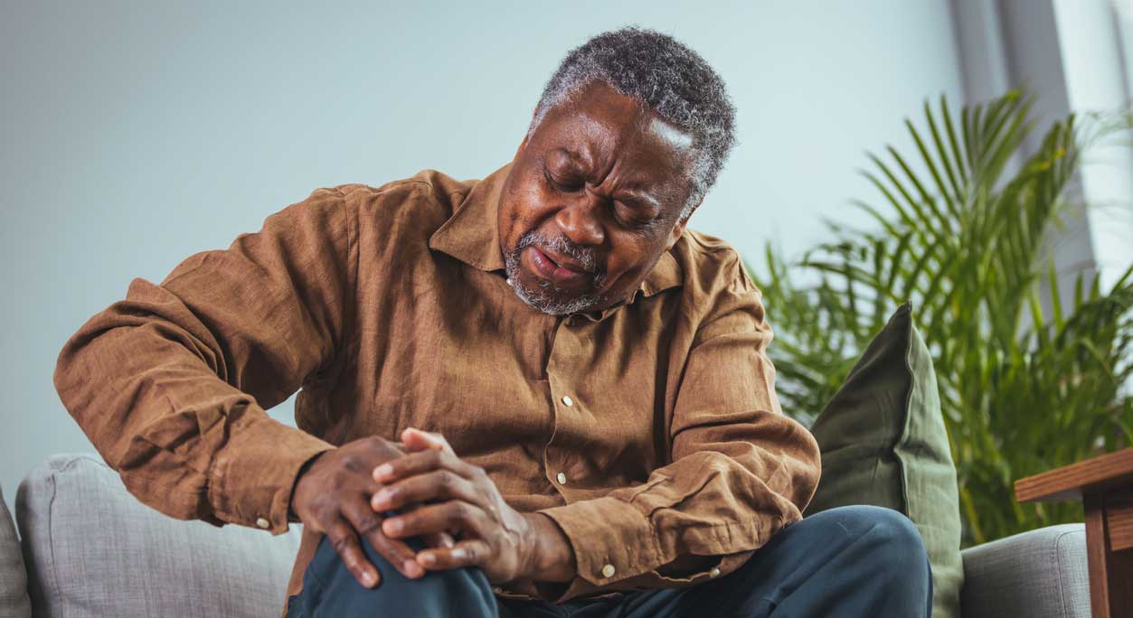 An older man sits in a chair, holding his knee in pain.
