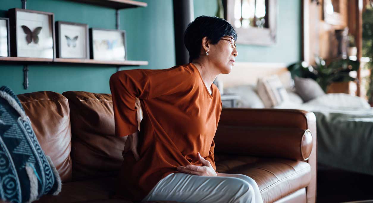 An older woman sits on a sofa, holding her back in pain.