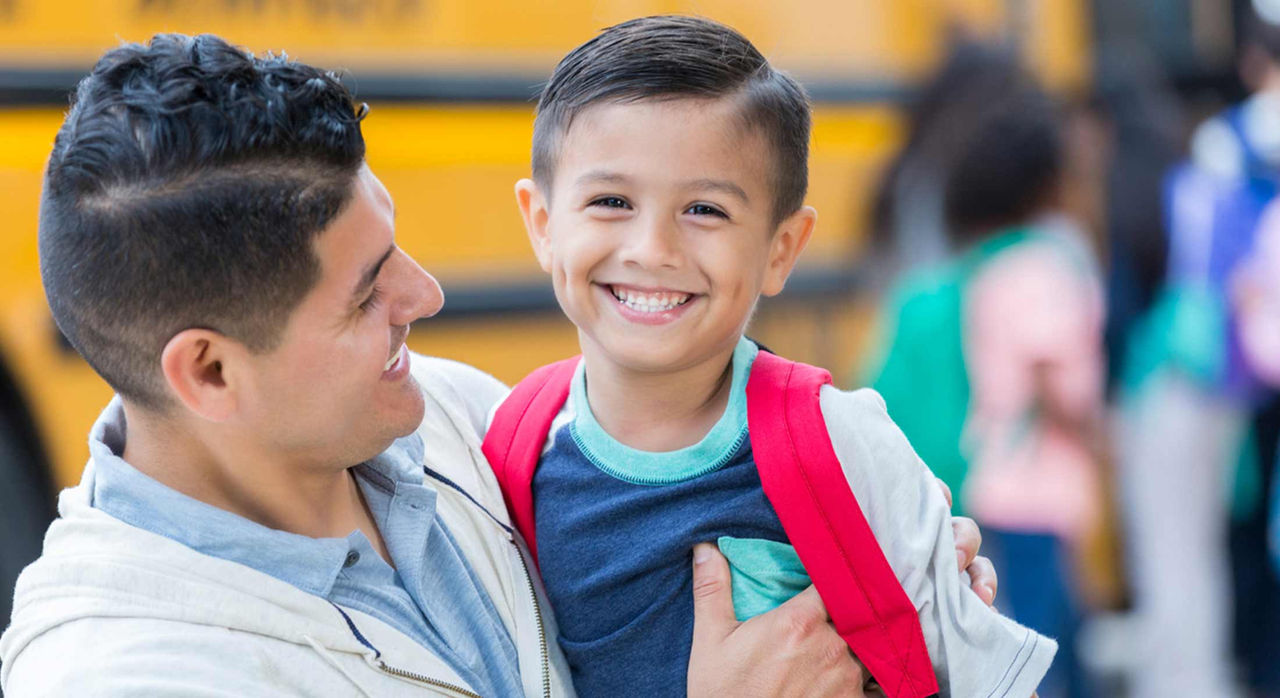 A father carrying his smiling child with a yellow school bus in the back