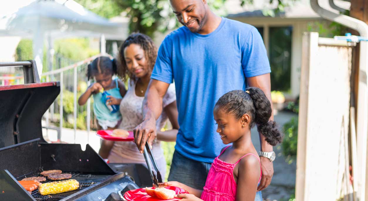 Young parents and their two little girls barbecue in their back yard.