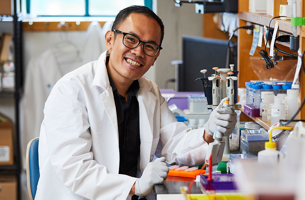 A smiling scientist piping liquid into tubes in a cluttered lab.