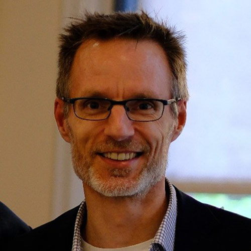 A man with short dark hair and short white facial hair is standing indoors. He is wearing glasses and a dark blazer over a light checkered shirt.