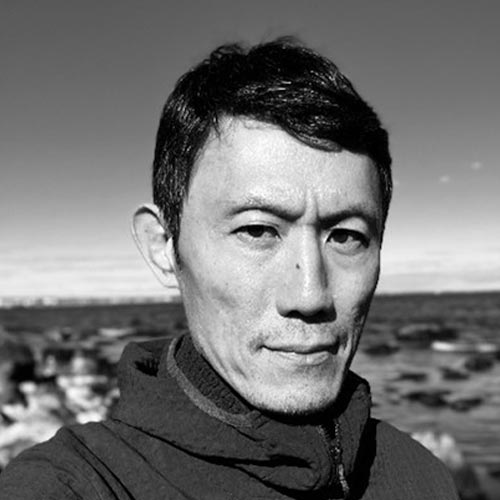 A person with black hair stands near a rocky shoreline in a hooded windbreaker.