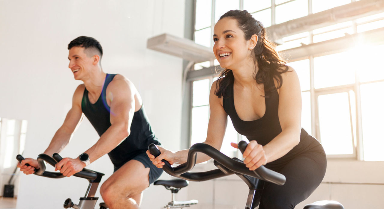 Two athletes use standing bikes for conditioning exercises in a room with natural light coming through