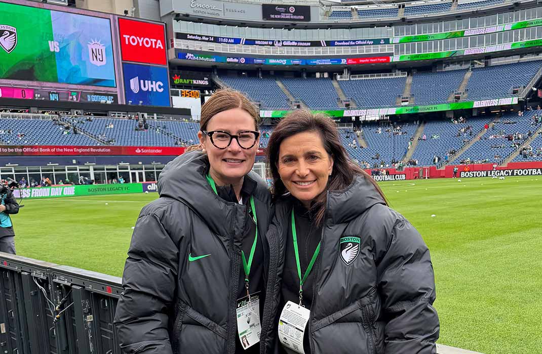 Two sports medicine doctors wearing black jackets with the Boston Legacy FC emblem while standing on the sideline of a soccer stadium.