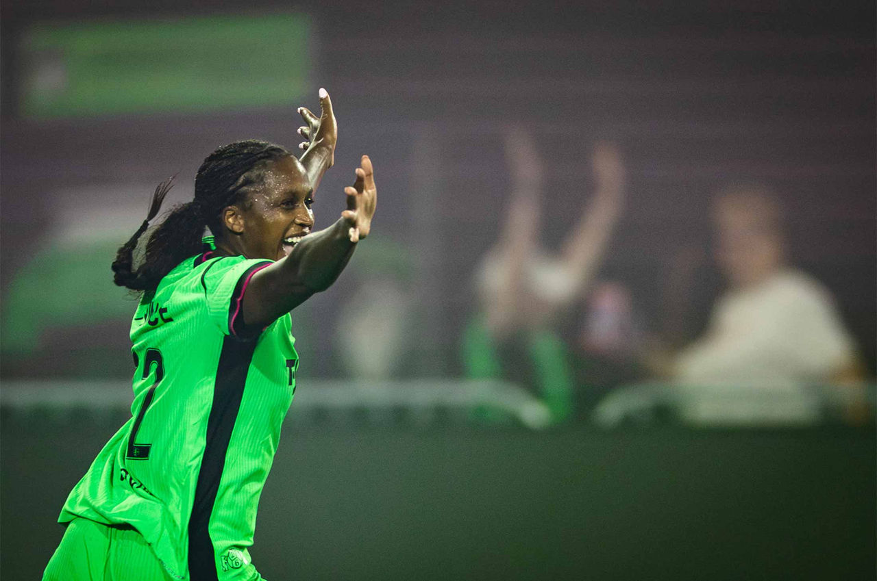 A women's soccer player in a bright green Boston Legacy FC uniform celebrates with arms raised during a match. She runs across the field, smiling and shouting in excitement. The background is blurred with spectators in the stands.