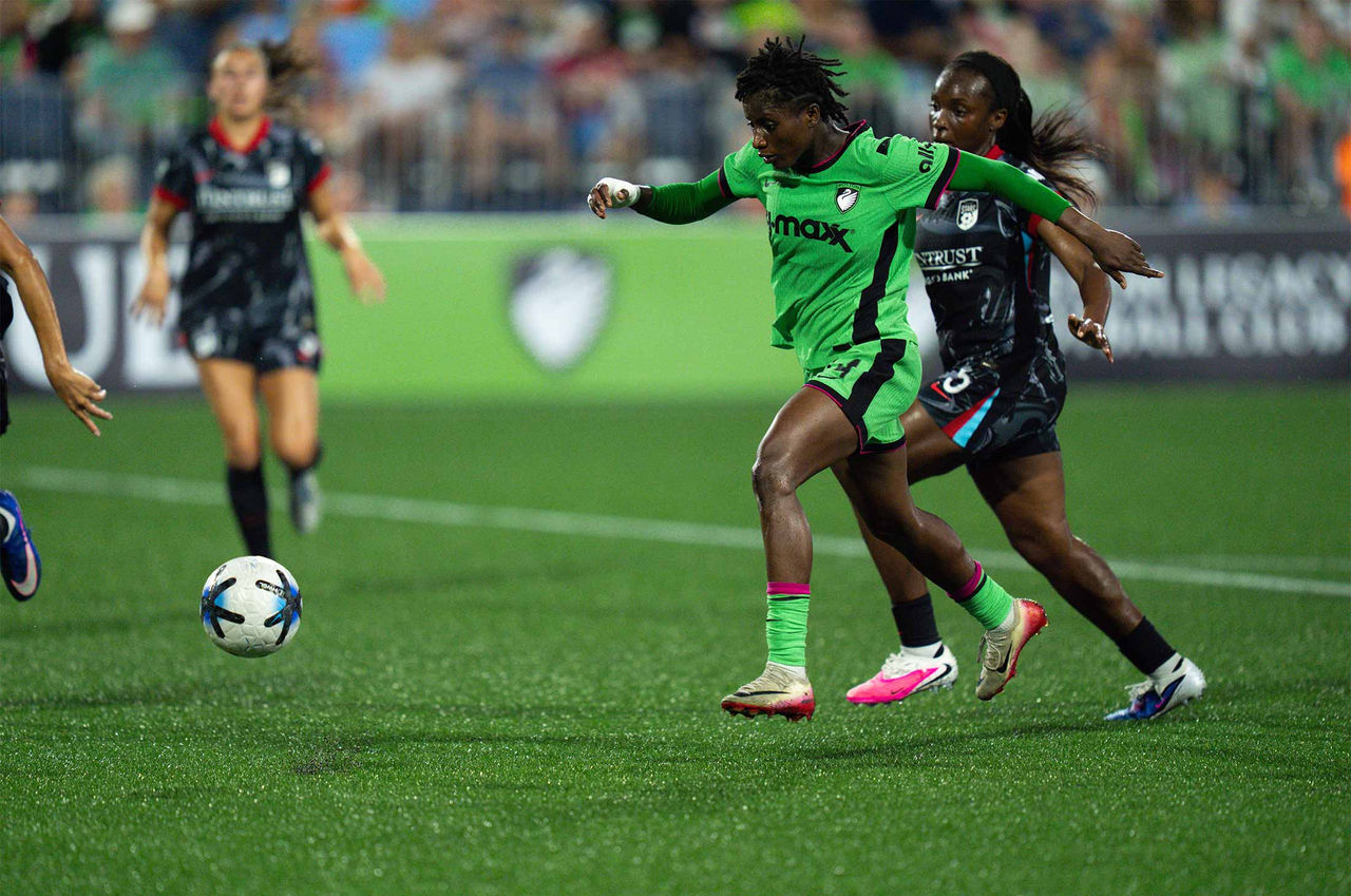 A women’s soccer match shows a Boston Legacy FC player in a bright green uniform sprinting with the ball while closely defended by an opponent in a dark kit from Chicago Stars FC. Other players are visible in the background on the field, with a crowd watching from the stands. The match takes place on a well-lit outdoor pitch under stadium lights.